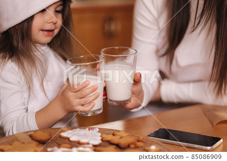 Portrait of beautiful tree year girl with her mom in santa hat sitting by the table in front of kitchen decorated fairy lights and drink fresh milk. Christmas mood Portrait of beautiful tree year girl with her mom in santa hat sitting by the table in front of kitchen decorated fairy lights and drink fresh milk. Christmas mood 85086909