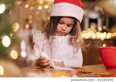 Little girl in santas hat decorates gingerbread using white glaze. Christmas and New Year traditions concept. Christmas bakery. Happy holidays Little girl in santas hat decorates gingerbread using white glaze. Christmas and New Year traditions concept. Christmas bakery. Happy holidays 85086949