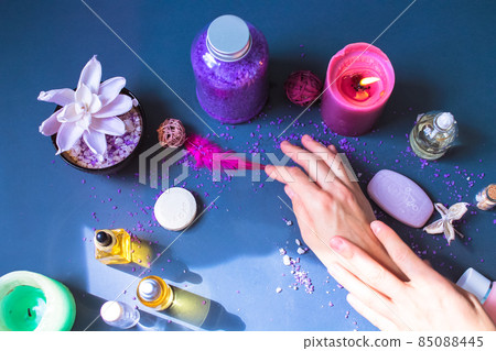Cosmetic products - bottles of facial and body oil, soap bars, purple bath salt, burning candle are lying on a dark grey table flat lay. Woman's hands view from above. Spa day. Female beauty concept. 85088445