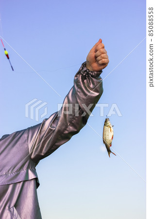 A woman's hand in a silver rain jacket holds a fishing line with a hooked fish in a body of water, pond, river, lake against blue sky. Recreation in nature at sunny day. Hunting and fishing on a rod. 85088558