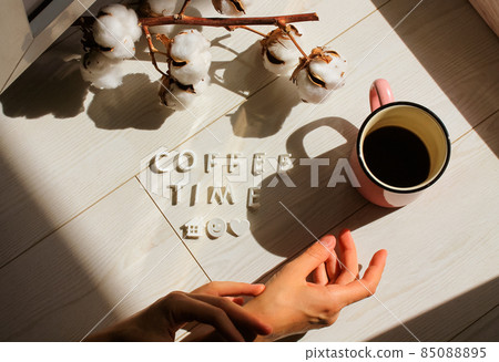 Tender woman's hands reaching for a pink cup of black coffee on a white wooden floor. Contrasting shadows, morning light. Cotton flower branch. Hot aromatic drink in a mug. Cozy homy atmosphere. Fall. 85088895