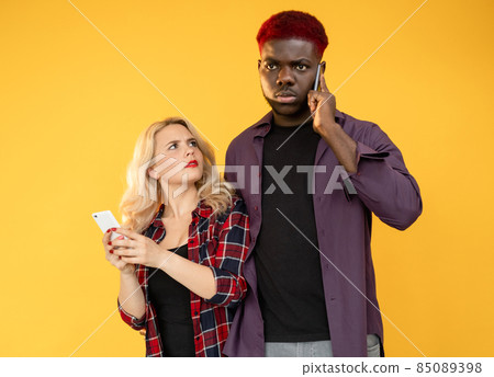 Phone call. Bad news. Unexpected problem. Booking cancellation. Multiethnic couple of confused man on cell conversation with suspicious woman isolated on orange background. 85089398