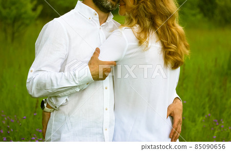 Newlyweds. Beautiful couple in love gently embracing standing in a green meadow in summer. Husband and wife in white shirts showing their feelings for each other. Devotion, purity, harmony, love theme 85090656