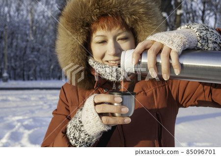 Smiling woman in red coat drinks hot tea from metal thermos. Leisure activity in winter forest at sunset. 85096701