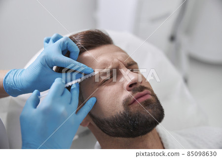 Ppatient grimaces of pain while careful cosmetologist does injection of lifting filler in his brow in clinic 85098630