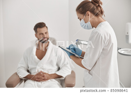 Administrator in mask and gloves fills form examining male patient in cosmetologycal clinic 85098641
