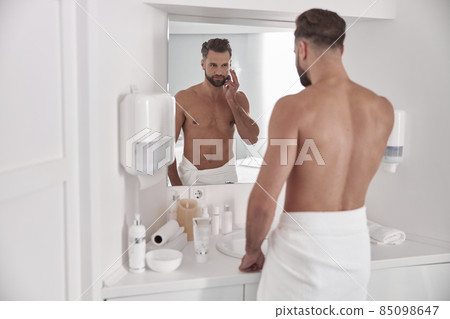 Attractive middle aged man applies cream onto face looking in mirror in bathroom Attractive middle aged man applies cream onto face looking in mirror in bathroom 85098647