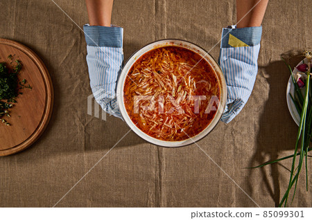 Top view of chef's hands in kitchen gloves, holding pot of freshly made borscht soup and placing it on table with linen tablecloth next to cutting board filled with chopped dill and green onions Top view of chef's hands in kitchen gloves, holding pot of freshly made borscht soup and placing it on table with linen tablecloth next to cutting board filled with chopped dill and green onions 85099301