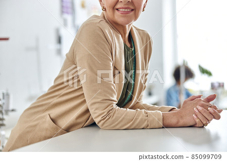 Smiling mature woman leans onto cutting table in spacious sewing workshop, closeup Smiling mature woman leans onto cutting table in spacious sewing workshop, closeup 85099709