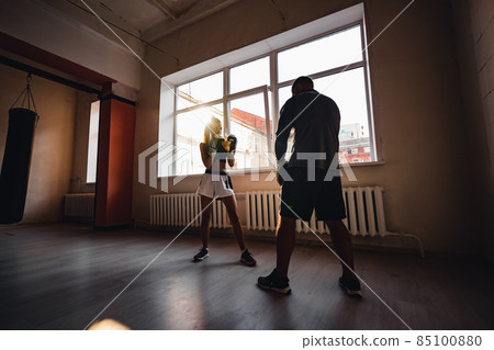 Medium shot of young woman doing boxing workout at the gym and punching her instructor, who is dodging her blows and controlling training process. 85100880