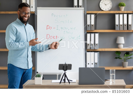 Cheerful millennial black guy teacher in glasses looks at device, shows on blackboard with english rules Cheerful millennial black guy teacher in glasses looks at device, shows on blackboard with english rules 85101603