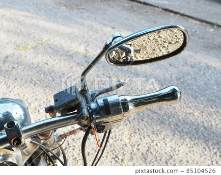Rearview mirror on a rust-coated motorcycle handlebar with reflective asphalt in the mirror 85104526