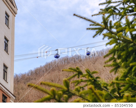 Two blue lifts ride a cable car against the backdrop of mountains with bare trees. View through the spruce branches 85104550