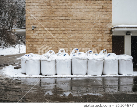 White bags of salt stand in the snow near a wet road against a wall with decorative brickwork. White bags of salt stand in the snow near a wet road against a wall with decorative brickwork. 85104677
