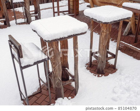 Tall wooden tables and bar stools of an outdoor cafe stand covered with snow in winter 85104870
