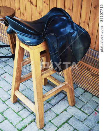 Wooden bar stool with a decorative leather saddle on the seat stands on the paving slabs near the wooden bar on the street Wooden bar stool with a decorative leather saddle on the seat stands on the paving slabs near the wooden bar on the street 85107716