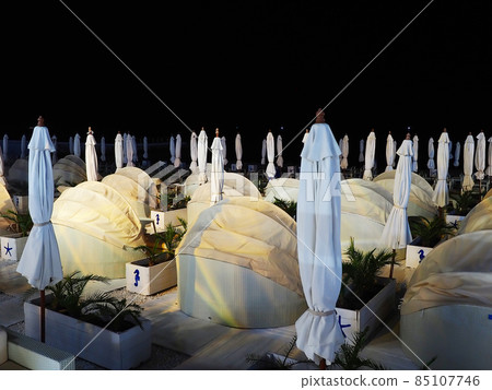 Night beach with many semicircular sofas with awnings and folded umbrellas against a dark sky Night beach with many semicircular sofas with awnings and folded umbrellas against a dark sky 85107746