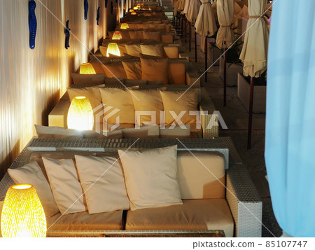 Light rattan sofas with cushions and lamps stand in a row in the semi-darkness. Beach luxury recreation area. Beach cafe 85107747