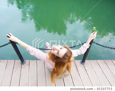 A smiling girl sits on a wooden bridge over the lake holding on to iron pillars. Top view A smiling girl sits on a wooden bridge over the lake holding on to iron pillars. Top view 85107753