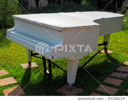 An old white wooden grand piano stands on a green lawn in the park. Park decor An old white wooden grand piano stands on a green lawn in the park. Park decor 85108224