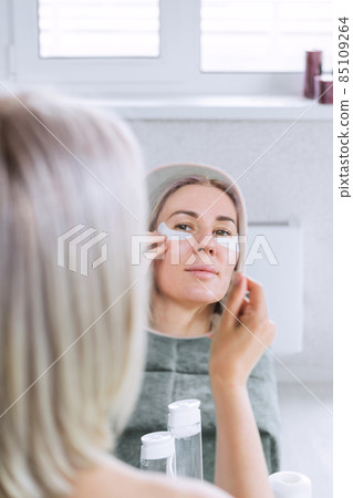 A beautiful young woman is taking care of her face, looking in the mirror applying eye patches. SPA. Facial care at home A beautiful young woman is taking care of her face, looking in the mirror applying eye patches. SPA. Facial care at home 85109264