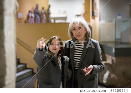 Aged female tutor with preteen girl exploring showpieces in museum 85109524