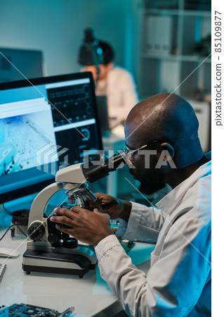 Young man of African ethnicity sitting by workplace and looking in microscope Young man of African ethnicity sitting by workplace and looking in microscope 85109877
