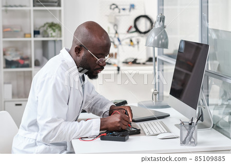 Serious young African repairman in whitecoat soldering circuit board 85109885