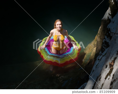 Smiling girl in a multicolored swimming circle stands in the water in the rays of the sun in a shady gorge of canyons Smiling girl in a multicolored swimming circle stands in the water in the rays of the sun in a shady gorge of canyons 85109909