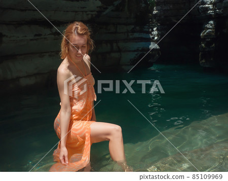 Sunlit smiling girl touching clear water while standing in shady rocky lagoon 85109969
