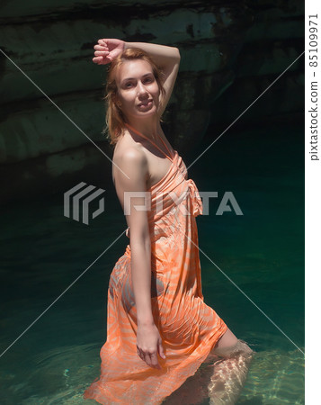 Smiling girl in orange sundress stands in the light of the sun in a rocky lagoon Smiling girl in orange sundress stands in the light of the sun in a rocky lagoon 85109971