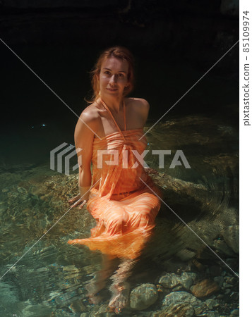 Smiling girl in orange sundress sits in clear river water in rocky lagoon 85109974