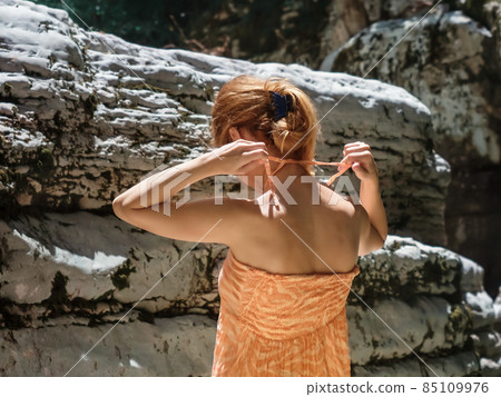 A girl ties the straps of a sundress while standing against the background of relief canyons under bright sun rays 85109976