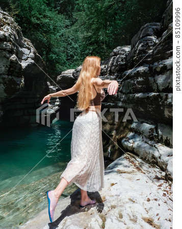 A longhaired girl stands in a swallow pose near a turquoise river in a rocky environment on a sunny day 85109996