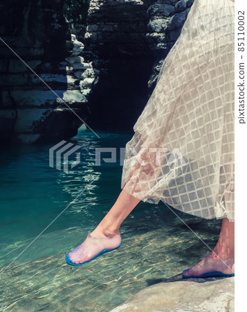 Female feet taking a step into clear turquoise water in a rocky environment 85110002