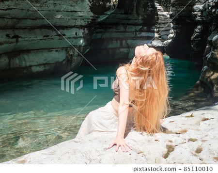 A longhaired girl sits with her head thrown back on the rocky bank of a turquoise river surrounded by canyons 85110010