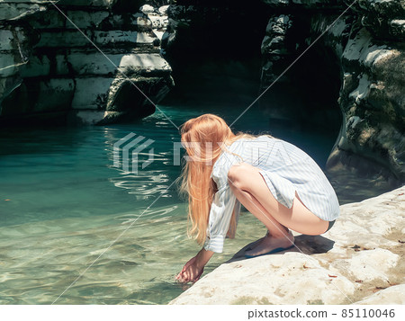 A longhaired girl in a shirt washes her hands in a turquoise pond while squatting on a rocky shore in a canyon gorge A longhaired girl in a shirt washes her hands in a turquoise pond while squatting on a rocky shore in a canyon gorge 85110046
