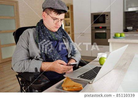 Young man in casualwear sitting in wheelchair while scrolling in smartphone 85110339