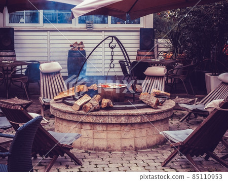 A cauldron of vegetables hangs over a stone well with wood and sun loungers around outdoors. Street cafe 85110953