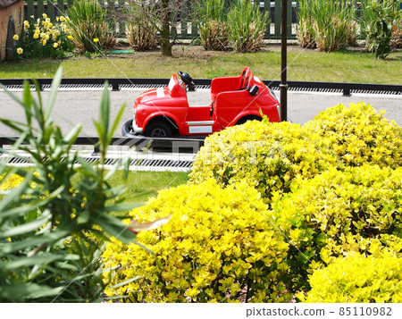 A small children's red car stands on a miniature road surrounded by bright bushes. Children's karting track in the park A small children's red car stands on a miniature road surrounded by bright bushes. Children's karting track in the park 85110982