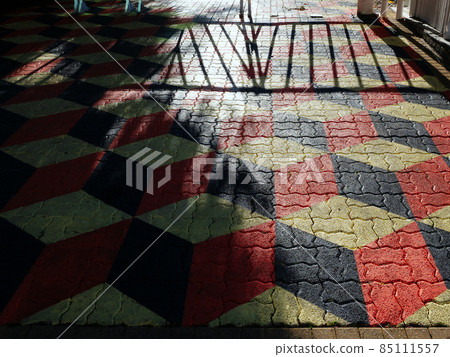 Terrain with paving slabs with a red-black geometric pattern and a falling shadow from trees and a fence 85111557