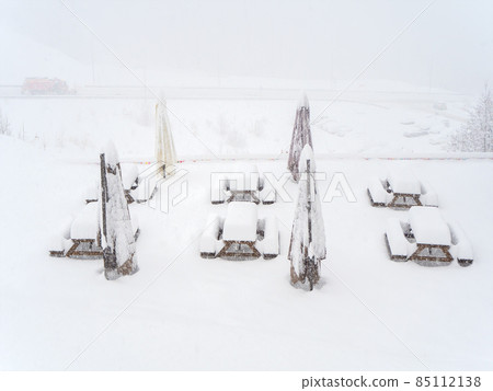 Snow-covered tables with benches and folded outdoor umbrellas stand on the site on a foggy snowy day Snow-covered tables with benches and folded outdoor umbrellas stand on the site on a foggy snowy day 85112138