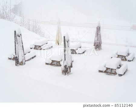 Snow-covered tables with benches and folded outdoor umbrellas stand on the site on a winter day during a blizzard 85112140