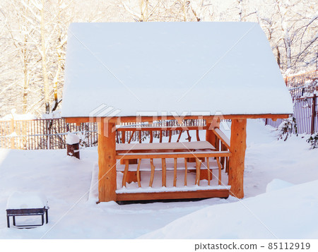 A wooden gazebo covered with snow stands on a snow-covered meadow among trees 85112919