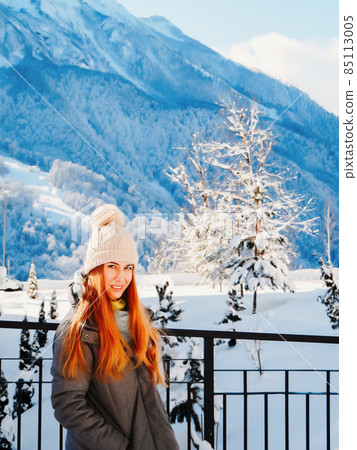 Redhaired smiling girl stands at the fence against the background of high snow-capped mountains Redhaired smiling girl stands at the fence against the background of high snow-capped mountains 85113005
