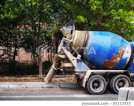A blue concrete mixer stands on the side of the road near the curb against the backdrop of a house and trees A blue concrete mixer stands on the side of the road near the curb against the backdrop of a house and trees 85113555