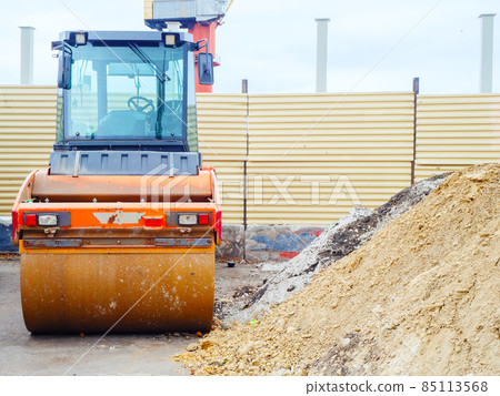 A roller to roll asphalt stands near a pile of sand against the background of a construction fence A roller to roll asphalt stands near a pile of sand against the background of a construction fence 85113568
