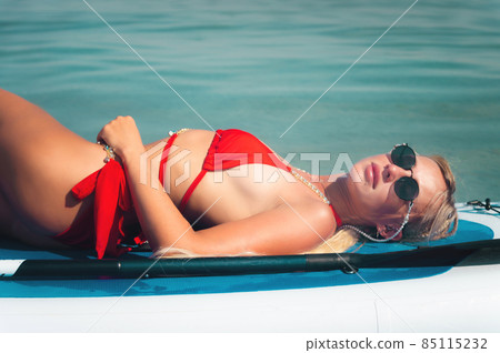 Young sexy surfer woman in red swimsuit and sunglasses lying on her sup board. Relax on stand up paddle on a sunny day Camping concept 85115232