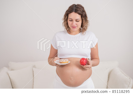 A pregnant woman is holding a red apple and donut. Choice of food. 85118295