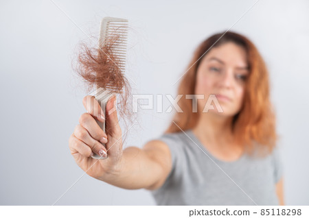 Caucasian woman with a grimace of horror holds a comb with a bun of hair. Hair loss and female alopecia. 85118298